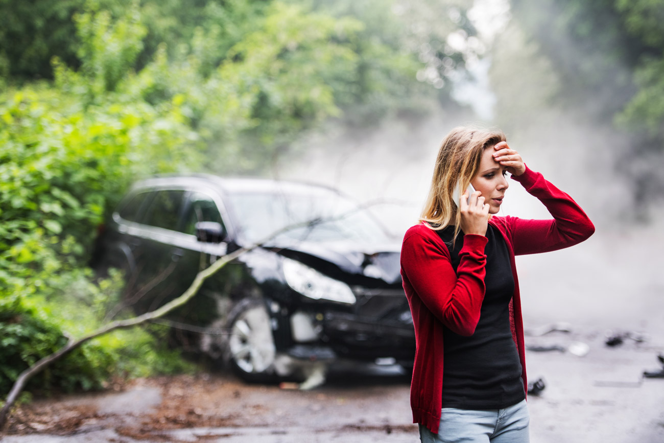 Woman on the phone after getting into a car accident with her damaged car in the background